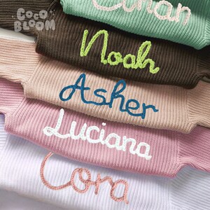 May include: A stack of five knitted sweaters with different names embroidered on them in various colors. The names are "Coma", "Luciana", "Asher", "Noah", and "Olivia".