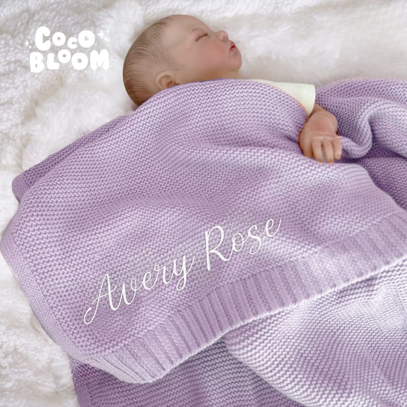 Op de afbeelding: Een lila gebreide babydeken met de naam "Avery Rose" in witte letters. De deken is gedrapeerd over een babypop op een witte, zachte ondergrond. Het Coco Bloom-logo staat in de linkerbovenhoek.