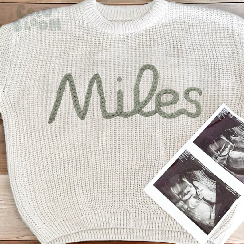 May include: A white knitted sweater with the name "Miles" embroidered in green yarn. Two black and white ultrasound images are in the background.