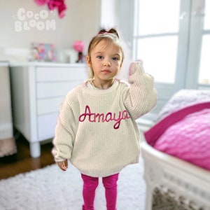 May include: A young girl wearing a white knitted sweater with the name "Amaya" embroidered in pink on the front. She is also wearing pink leggings.