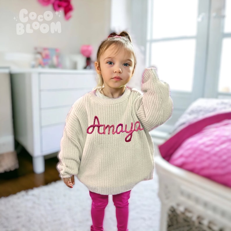 May include: A young girl wearing a white knitted sweater with the name "Amaya" embroidered in pink on the front. She is also wearing pink leggings.