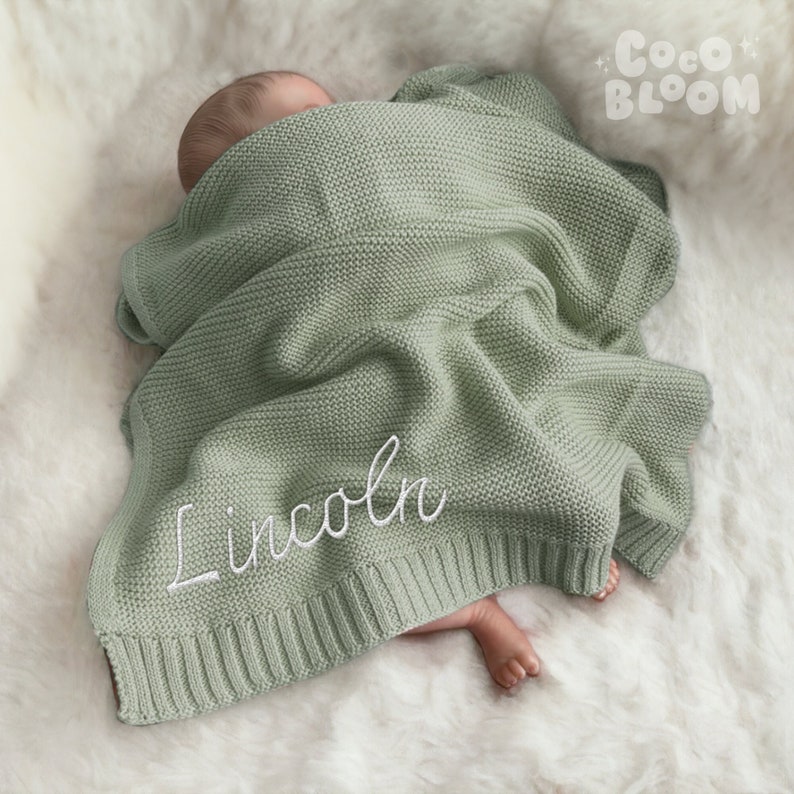 Op de afbeelding: Een saliegroene gebreide babydeken met de naam "Lincoln" geborduurd in wit script. De deken is gedrapeerd over een baby die op een wit, zacht oppervlak ligt. Het Coco Bloom-logo is rechtsboven zichtbaar.