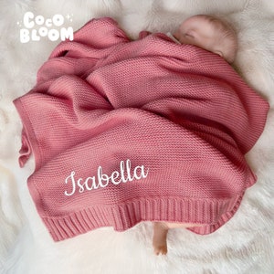 Op de afbeelding: Een roze gebreide babydeken met de naam "Isabella" in witte letters geborduurd. De deken is om een slapende baby gewikkeld. Het Coco Bloom-logo staat in de linkerbovenhoek.