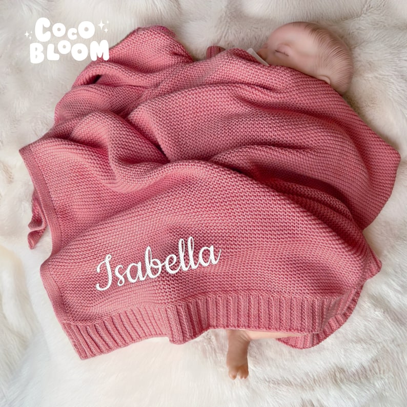 Op de afbeelding: Een roze gebreide babydeken met de naam "Isabella" in witte letters geborduurd. De deken is om een slapende baby gewikkeld. Het Coco Bloom-logo staat in de linkerbovenhoek.