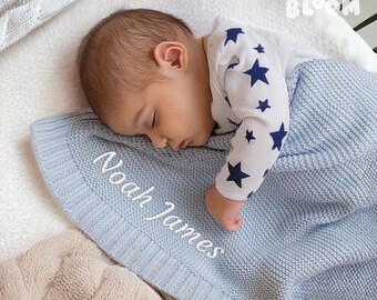 May include: A light blue knitted baby blanket with the name "Noah James" embroidered in white. The blanket is folded and a baby is sleeping on top of it. The baby is wearing a white onesie with blue stars.
