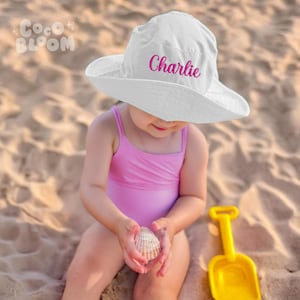 Peut inclure: Un chapeau de soleil blanc à large bord avec le nom "Charlie" brodé en rose sur le devant. Un jeune enfant porte le chapeau et tient un coquillage.