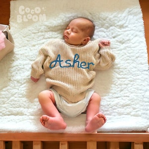 May include: A baby wearing a beige knitted sweater with the name "Asher" embroidered in blue. The baby is lying on a white fluffy blanket.