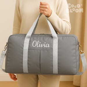 May include: A grey duffel bag with the name "Olivia" embroidered in white. The bag features a shoulder strap and two handles. The bag is made of a durable material, suitable for travel or everyday use. It has a zip closure.