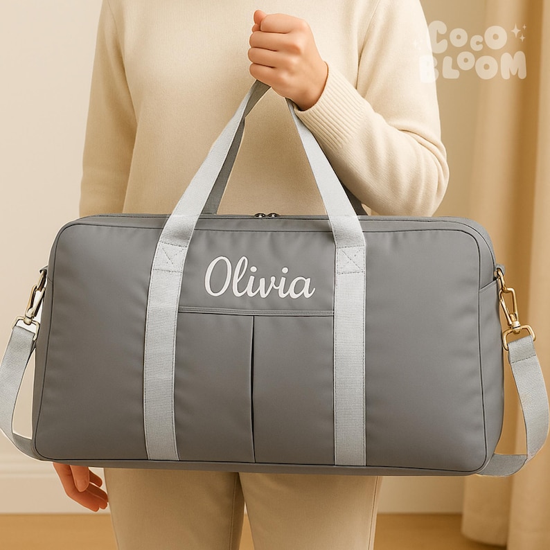 May include: A grey duffel bag with the name "Olivia" embroidered in white. The bag features a shoulder strap and two handles. The bag is made of a durable material, suitable for travel or everyday use. It has a zip closure.