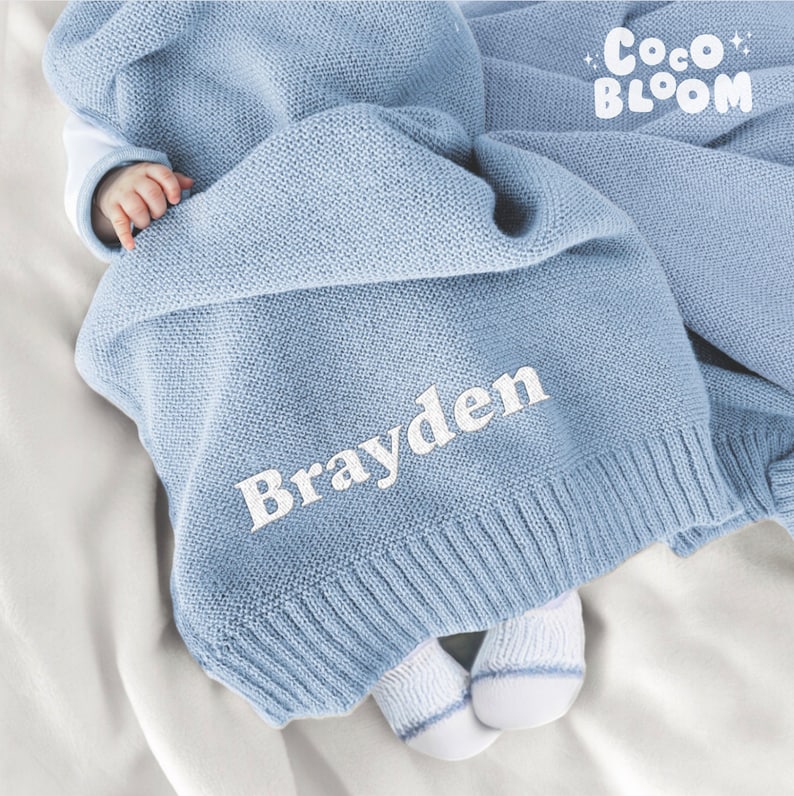 Op de afbeelding: Een lichtblauwe gebreide babydeken met de naam "Brayden" in het wit geborduurd. De deken is over een baby gedrapeerd, met het "Coco Bloom" logo rechtsboven. De deken is gemaakt van zacht materiaal.