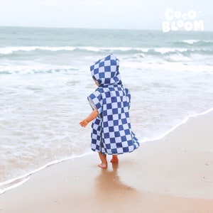 Peut inclure: Un enfant portant une serviette &agrave; capuche &agrave; carreaux bleus et blancs sur une plage de sable avec des vagues oc&eacute;aniques en arri&egrave;re-plan. La serviette porte le texte "Coco Bloom".