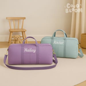 May include: Two personalised duffel bags. One is lavender with "Hailey" in white script, the other is light blue with "Xavier". Both have handles, shoulder straps, and gold-coloured hardware. A small wooden chair and a teddy bear are in the background.