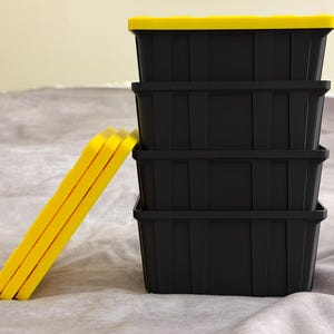 May include: Four black storage boxes with yellow lids stacked together. Three yellow lids are propped against the side. The boxes are on a grey surface.