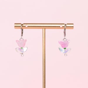 May include: A pair of pink flower earrings with iridescent accents hanging from a gold earring stand.