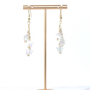 Op de afbeelding: Een paar bungelende oorbellen met een hemels thema. Elke oorbel heeft een goudkleurige haak en ketting, die een cluster van iriserende stervormige kralen ophangt. De oorbellen worden getoond op een goudkleurige sieradenstandaard tegen een witte achtergrond.