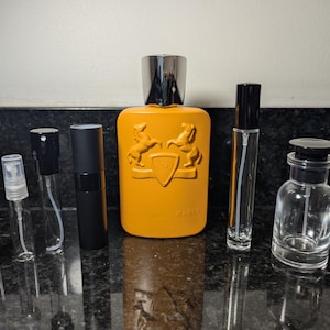 May include: A collection of perfume bottles, including a large orange bottle with a black and gold cap, a smaller clear bottle with a black cap, and several smaller clear bottles with black and silver caps. The bottles are arranged on a black surface.