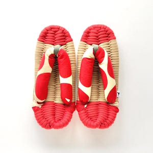 以下が含まれることがあります： 伝統的な日本の下駄のペア。サンダルは、赤と白の模様のストラップが付いた織りのベージュのベースが特徴です。つま先部分とベースの端は赤色です。サンダルは屋外での使用向けに設計されています。