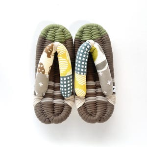 May include: A pair of traditional Japanese geta sandals. The sandals feature a woven brown base with a green top and patterned fabric straps in yellow, gray, and white. The geta are designed for outdoor wear.