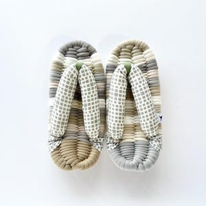 May include: A pair of traditional Japanese geta sandals. The sandals feature a woven platform in shades of gray, beige, and white. The thong straps are light gray with a white grid pattern. A small green accent is at the front.