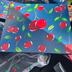 May include: A square wooden tray painted in a deep blue colour featuring a hand-painted pattern of bright red cherries with green leaves and brown stems scattered across the surface. The tray has a smooth finish and a flat, rectangular design.