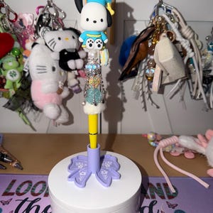 Puede incluir: Un bolígrafo decorativo con un personaje de dibujos animados que lleva un birrete. El bolígrafo amarillo tiene un adorno de joyas y se asienta en una base blanca con un diseño de lazo morado. Otros accesorios son visibles en el fondo.