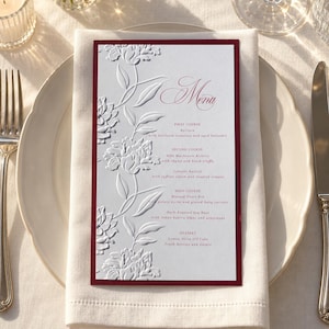 May include: A rectangular menu card with a deep red border and embossed white floral patterns on the left side. The text reads Menu, First Course, Second Course, Main Course, and Dessert, listing various food items in a formal serif font.