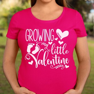 May include: A magenta maternity t-shirt with the text "GROWING my little Valentine" in white script. The design includes a heart, baby footprints, and small hearts. The shirt has a scoop neck and short sleeves.