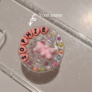 May include: Clear phone case with a circular, decorative element. The element features a pink teddy bear, heart-shaped beads spelling "SOPHIE", and colorful embellishments. The case is filled with glitter.