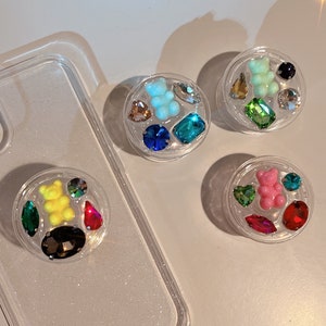 May include: Three clear phone grips with colorful gummy bear charms and rhinestones. Each grip has a different color gummy bear: pink, green, and blue.
