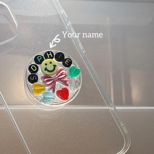 May include: A clear phone case with a pop socket featuring a smiley face, a pink bow, and colorful hearts. The letters "SOPHIE" are spelled out around the smiley face.