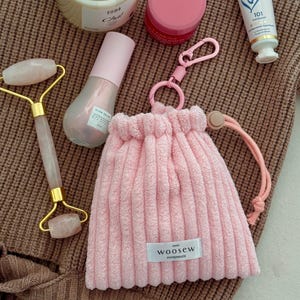 Könnte beinhalten: Eine Auswahl an Schönheits- und Pflegeprodukten, darunter ein rosa Kordelzugbeutel mit dem Etikett "WOOSEW HANDMADE", eine Gesichtsrolle aus Rosenquarz, eine Kerze, Lippenbalsam und eine Tube Lanolips 101 Salbe. Auf einer braunen Strickoberfläche.