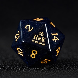 May include: A dark blue, glittery twenty-sided die featuring gold-coloured numbers. One face displays the text H&K, a graphic of two interlocking rings, and the date 10.06.2026. The die has a smooth, polished finish with sharp, defined edges.