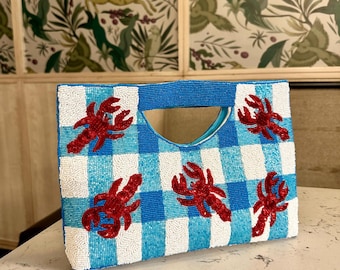 May include: A beaded handbag with a blue and white gingham pattern. The bag features red lobster appliqués and a blue beaded handle. The background is a light-colored surface with a patterned wallpaper.