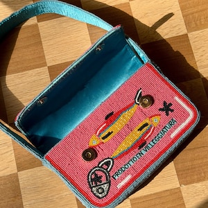 May include: A beaded pink handbag with a blue handle and interior. The bag features a design of two yellow and orange fish, a padlock, and the text "PRODOTTO EN VILLEGGIATURA" in white. The bag is open, revealing the blue interior.