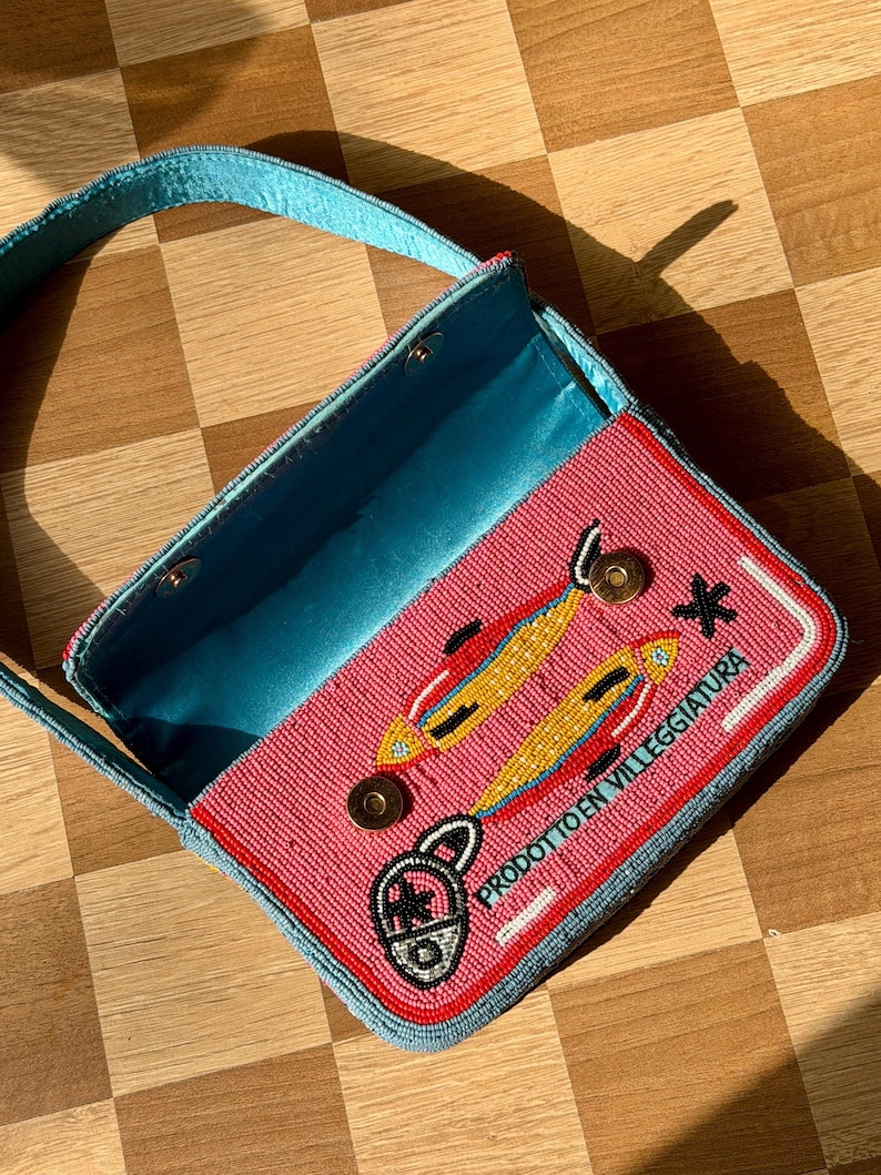 May include: A beaded pink handbag with a blue handle and interior. The bag features a design of two yellow and orange fish, a padlock, and the text "PRODOTTO EN VILLEGGIATURA" in white. The bag is open, revealing the blue interior.
