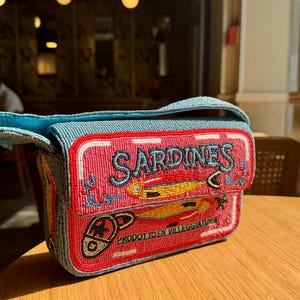 May include: A beaded handbag with a design resembling a sardine can. The bag features a pink front with the word "SARDINES" in blue, and illustrations of sardines. The bag has a blue strap and a red border.