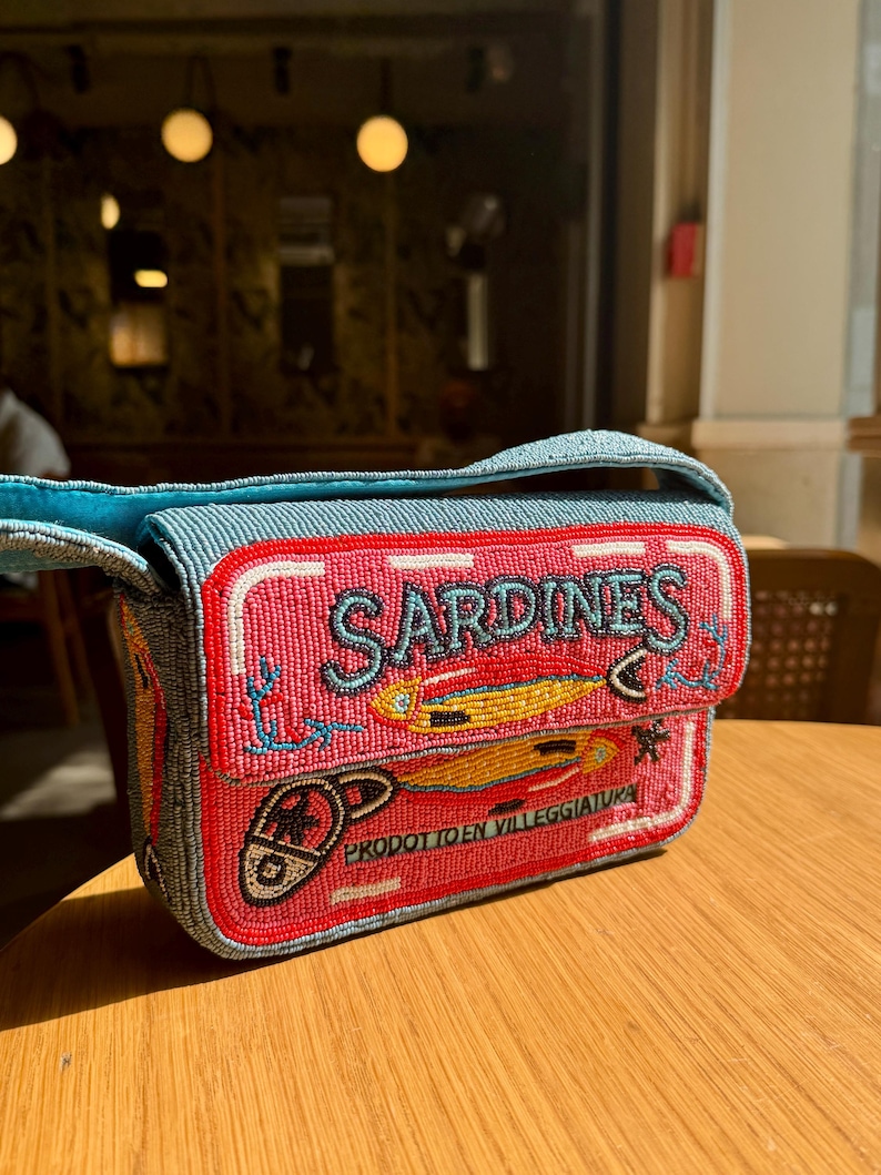 May include: A beaded handbag with a design resembling a sardine can. The bag features a pink front with the word "SARDINES" in blue, and illustrations of sardines. The bag has a blue strap and a red border.