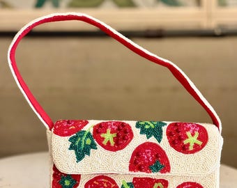 Puede incluir: Bolso de mano de cuentas color crema con asa y solapa rojas. El bolso está decorado con diseños de tomates rojos y hojas verdes. El asa es roja con ribete blanco.