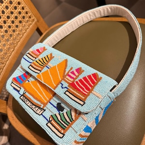 May include: A light blue beaded handbag with a white handle. The bag features a design of colorful sailboats and the word "NANTUCKET" in a rectangular label. The bag is resting on a green chair.