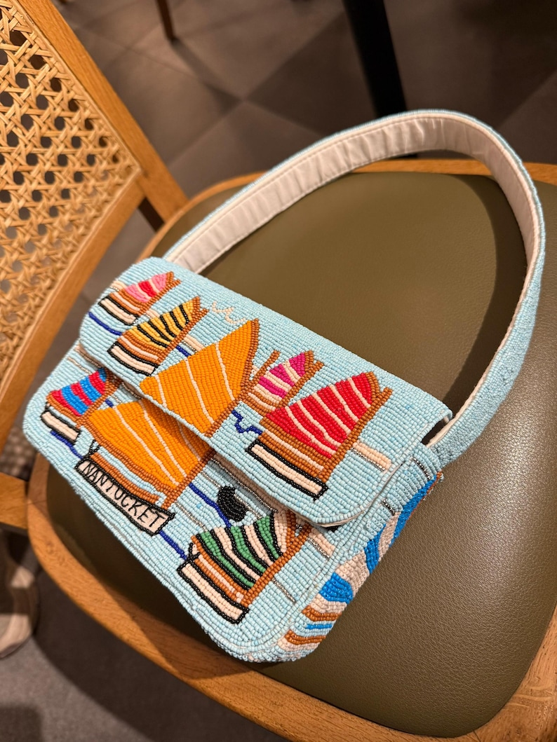 May include: A light blue beaded handbag with a white handle. The bag features a design of colorful sailboats and the word "NANTUCKET" in a rectangular label. The bag is resting on a green chair.