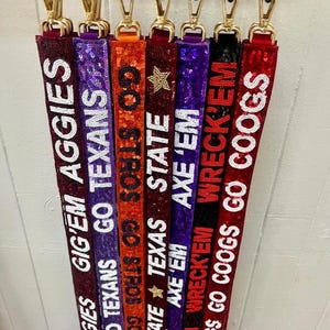 May include: A collection of beaded lanyards in various colors, each featuring text and a gold-colored clasp. The lanyards display phrases like "GIG'EM AGGIES", "GO TEXANS", and "TEXAS STATE". The beads are white, and the text is in a bold, clear font.