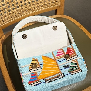 May include: A light blue beaded handbag with a sailboat design. The bag features an open top with a white interior and a handle. The sailboat design includes orange, red, pink, and green sails, with the word "NANTUCKET" displayed on the bag.