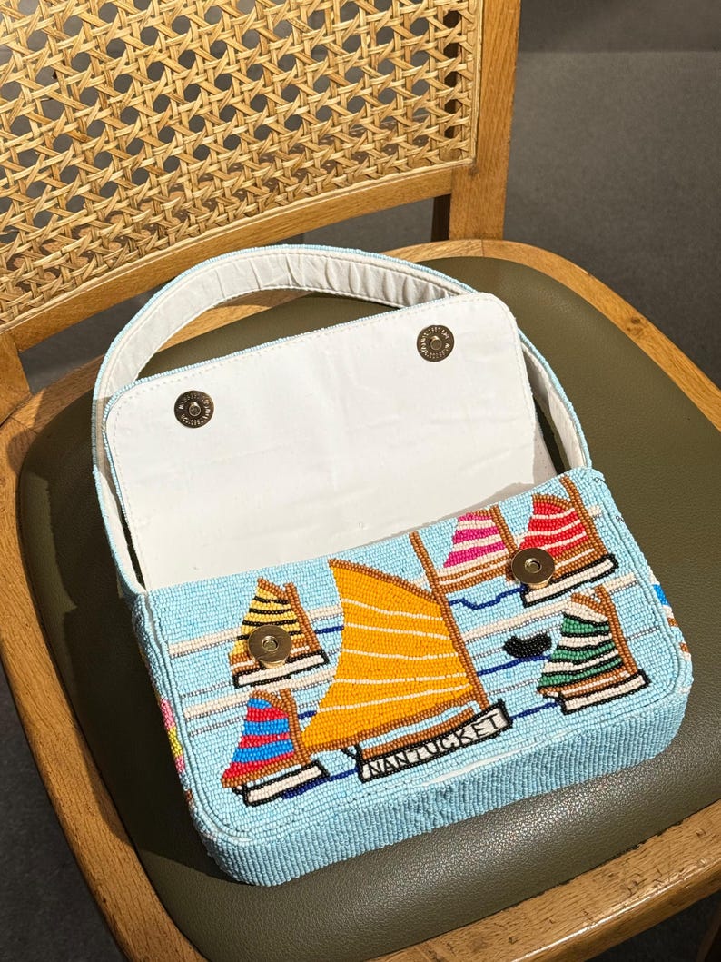 May include: A light blue beaded handbag with a sailboat design. The bag features an open top with a white interior and a handle. The sailboat design includes orange, red, pink, and green sails, with the word "NANTUCKET" displayed on the bag.