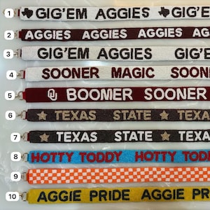 May include: A selection of beaded lanyards with different text and designs. The lanyards display phrases such as "GIG'EM AGGIES", "SOONER MAGIC", and "HOTTY TODDY". Colours include white, maroon, black, gold, blue, orange, and yellow.
