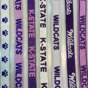 May include: Close-up of several beaded ribbons with text and paw prints. The ribbons feature the words "WILDCATS" and "K-STATE" in purple, white, and lavender. Some ribbons have paw print designs.