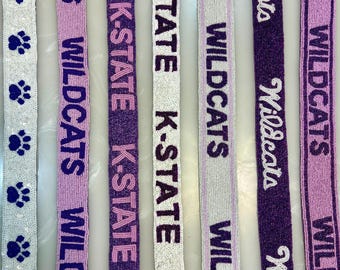 Może przedstawiać: Zbliżenie kilku koralikowych wstążek z tekstem i śladami łap. Wstążki zawierają słowa "WILDCATS" i "K-STATE" w kolorze fioletowym, białym i lawendowym. Niektóre wstążki mają wzory śladów łap.
