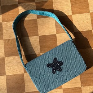 May include: A teal beaded handbag with a matching strap. The rectangular bag features a dark blue beaded starfish design on the front. The bag is set against a checkered wooden surface.