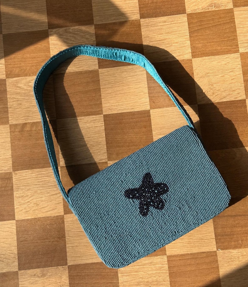 May include: A teal beaded handbag with a matching strap. The rectangular bag features a dark blue beaded starfish design on the front. The bag is set against a checkered wooden surface.
