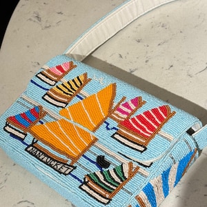 May include: A light blue beaded handbag with a white strap. The bag features a pattern of colorful boat designs, including orange, red, and blue, with the word "NANTUCKET" on the front. The bag has a rectangular shape.