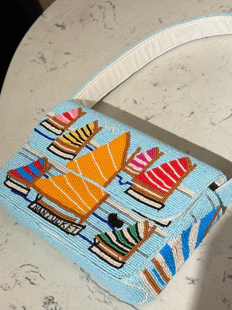 May include: A light blue beaded handbag with a white strap. The bag features a pattern of colorful boat designs, including orange, red, and blue, with the word "NANTUCKET" on the front. The bag has a rectangular shape.